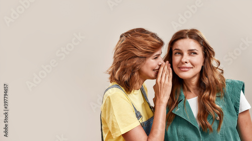 Close up portrait of two attractive young girls, twin sisters in casual wear telling secrets, gossiping, standing isolated over light background photo