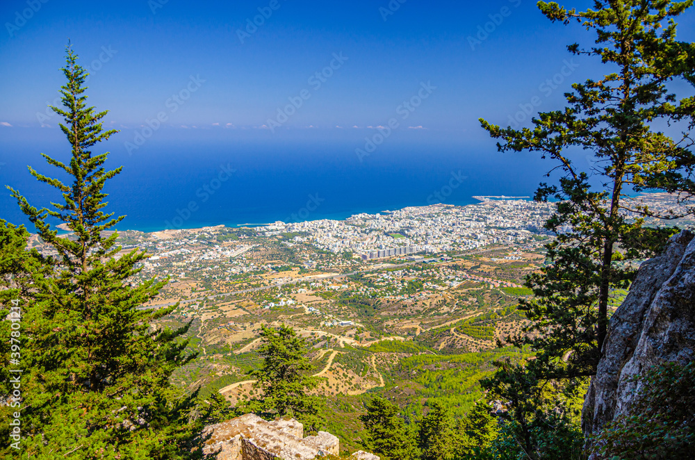 Aerial view of Kyrenia District valley with Kyrenia Girne city and Mediterranean sea in sunny day, green trees on rock of Kyrenia Girne mountains foreground, blue sky background, Northern Cyprus