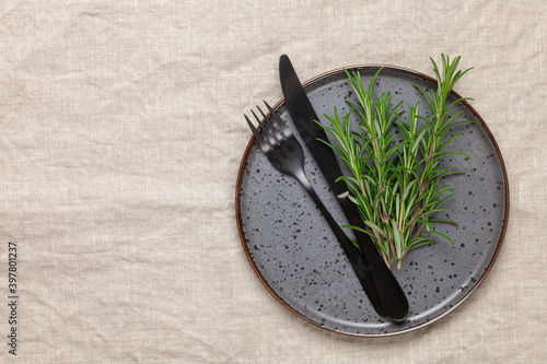 Black plate, cutlery and napkin with a sprig of rosemary on textile table. Top view. Table setting.