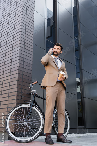 low angle view of businessman talking on smartphone and holding paper cup near bicycle and building