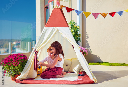 two happy kids playing together in teepee tent on sunny rofftop patio at home photo