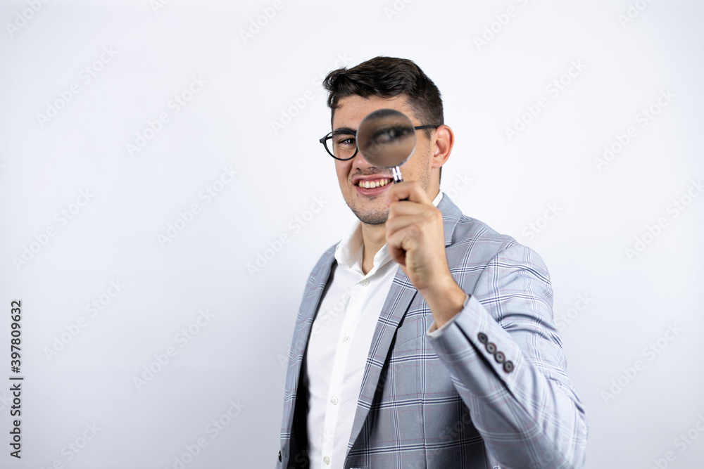 Business young man wearing a casual shirt over white background surprised looking through a magnifying glass Business young man wearing a casual shirt over white background surprised looking through a magnifying glass