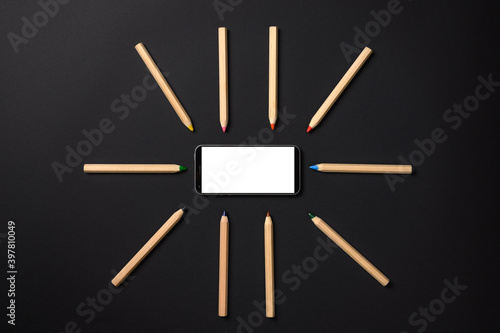 Pencils lie around the smartphone on a black background. Smartphone with blank white display