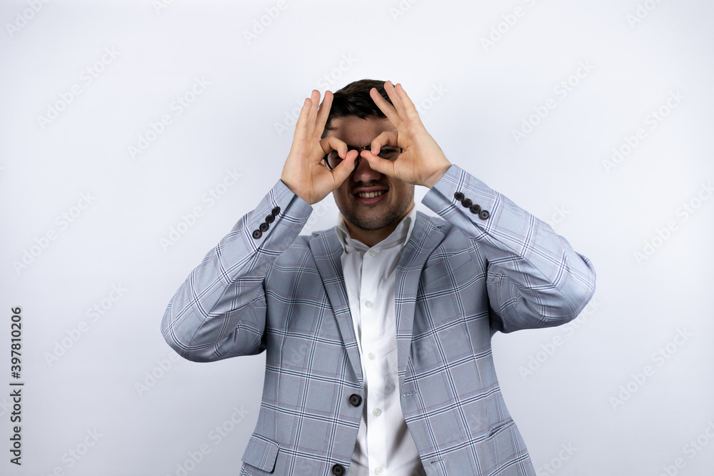 Business young man wearing a casual shirt over white background doing ok gesture shocked with smiling face, eye looking through fingers Business young man wearing a casual shirt over white background doing ok gesture shocked with smiling face, eye looking through fingers