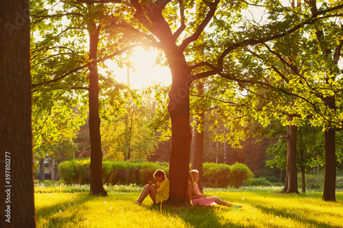 Two offended little girls sitting on grass leaning on tree trunk from opposite sides and sulking after quarrel in sunny summer park