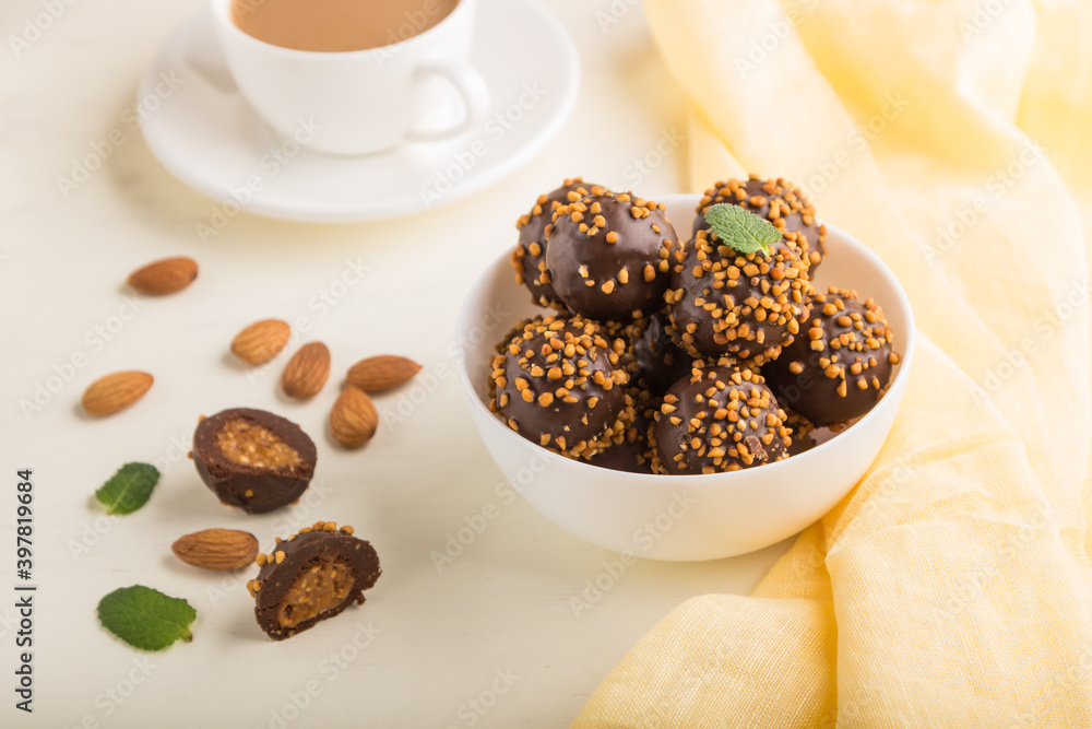 Chocolate caramel ball candies with almonds and a cup of coffee on a white wooden background. Side view, selective focus. Chocolate caramel ball candies with almonds and a cup of coffee on a white wooden background. Side view, selective focus.