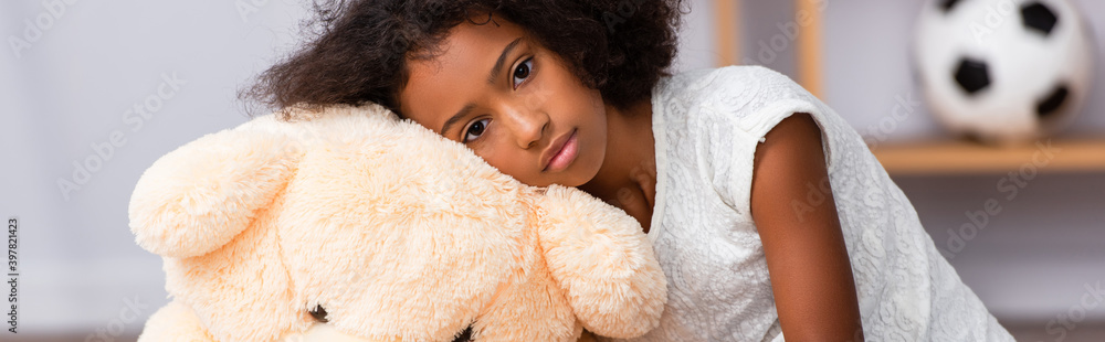 Upset african american girl looking at camera while leaning on teddy bear on blurred background, banner Upset african american girl looking at camera while leaning on teddy bear on blurred background, banner