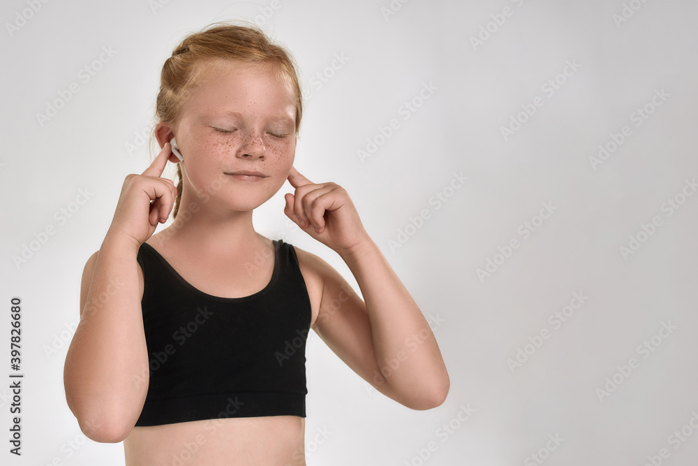Pretty little red haired sportive girl smiling, adjusting earphones while posing, standing with eyes closed isolated over white background