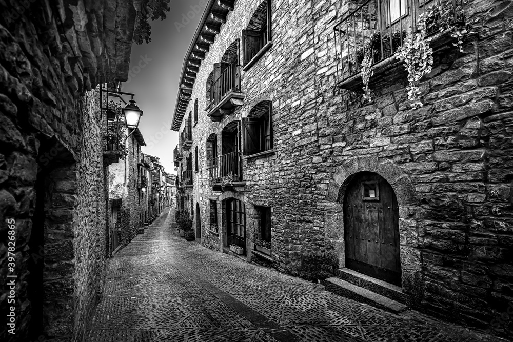 Black and white medieval old town street with stone houses and cobblestone floor, street lamps and dark and night atmosphere.
