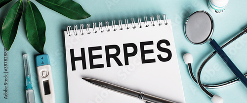 In a white notebook on a blue background, near a sheet of shefflers, a stethoscope, a syringe and an electronic thermometer, the word HERPES is written. Medical concept photo
