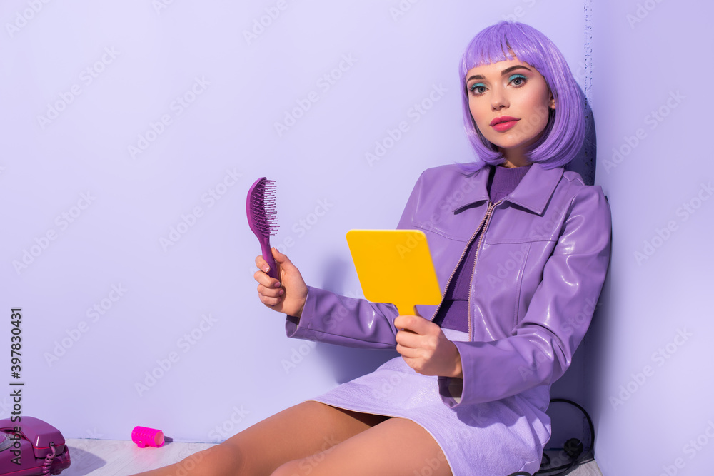 young woman dressed in doll style sitting with hair brush and mirror on violet colorful background