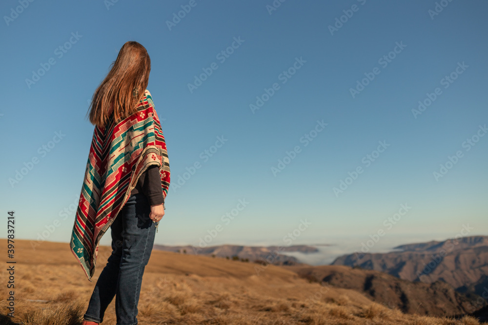 a girl in a colored national poncho stands and looks into the distance. girl on the background of the Caucasian mountains in full growth