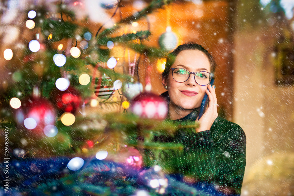 Portrait shot of smiling young woman using her mobile phone and talking with somebody while standing indoor behind the lighted window next to the christmas tree. Christmas time. Portrait shot of smiling young woman using her mobile phone and talking with somebody while standing indoor behind the lighted window next to the christmas tree. Christmas time.