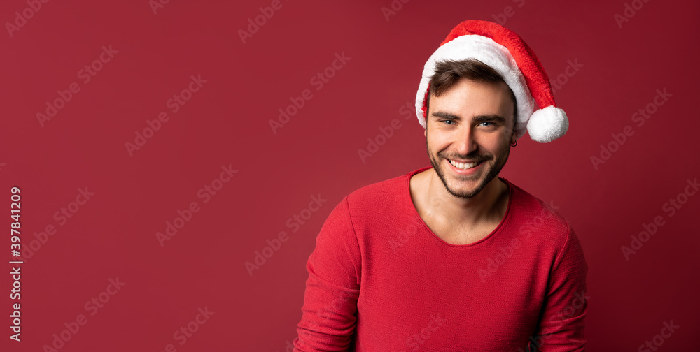 Young handsome caucasian guy in red sweater and Santa hats stands on red background in studio and teeth smiling Young handsome caucasian guy in red sweater and Santa hats stands on red background in studio and teeth smiling