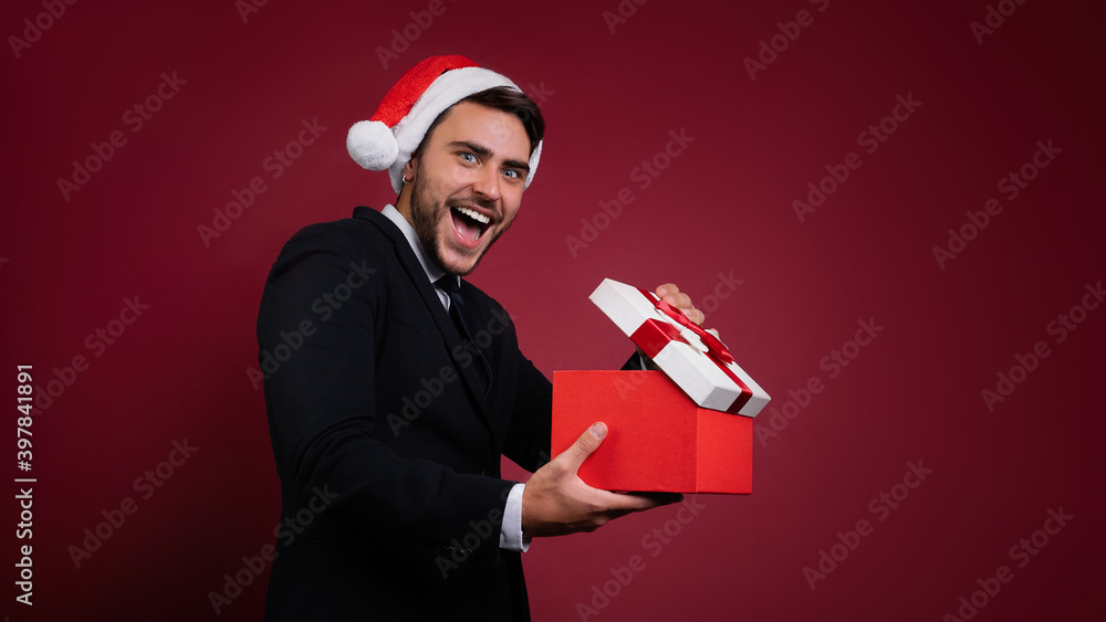 Young handsome caucasian guy in business suit and Santa hats stands on red background in studio and teeth smilie Holding red gift box in hand. Young handsome caucasian guy in business suit and Santa hats stands on red background in studio and teeth smilie Holding red gift box in hand.