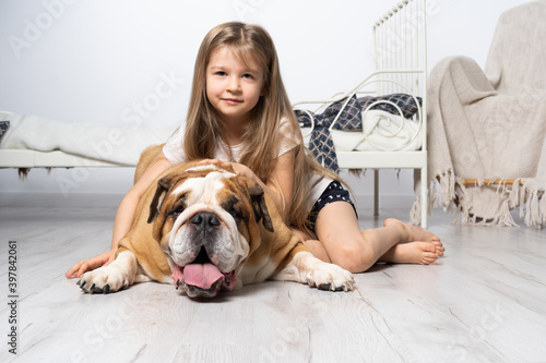 A young girl in a bedroom, sitting on the floor with her dog and stroking him. Child and dog. The English Bulldog is a purebred dog with a pedigree.