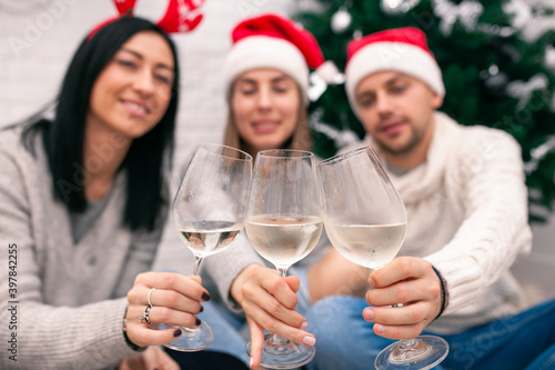 Young happy couple in Christmas hats near a Christmas tree kissing, holding glasses of wine. New Year celebration