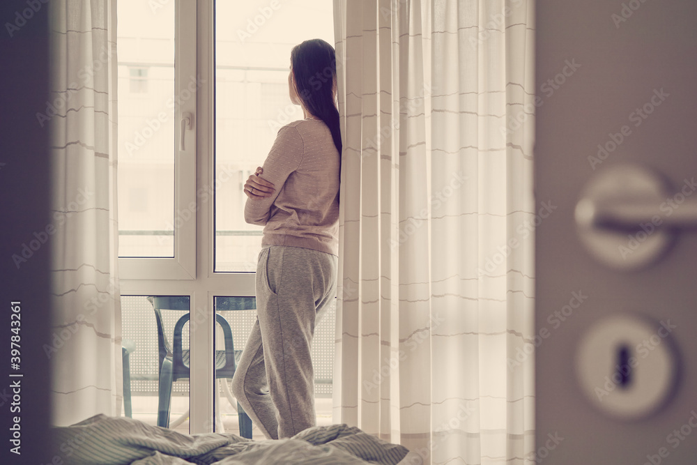 Woman looking out of a bedroom window with a messy bed Woman looking out of a bedroom window with a messy bed