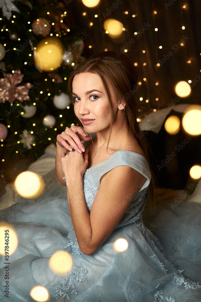 beautiful woman in a blue evening dress sits near a Christmas tree and dreams on the background of a garland. Winter holidays concept, new year, christmas, discounts, sales, advertising of  decoration