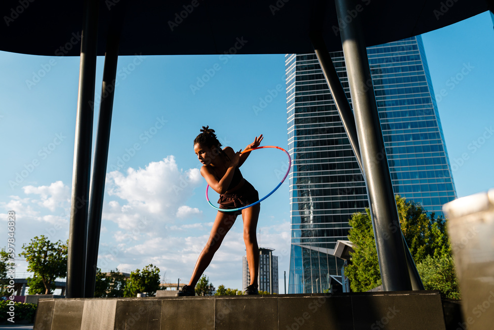 © VISTA by Westend61 - Sporty woman exercising with plastic hoops in modern city against blue sky © VISTA by Westend61 - Sporty woman exercising with plastic hoops in modern city against blue sky