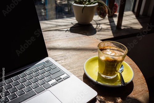 A cup (glass) of hot cafe latte coffee on yellow plate with stainless spoon put beside of a laptop computer notebook on wooden table in a coffee shop. Coffee break, work anywhere concept.
