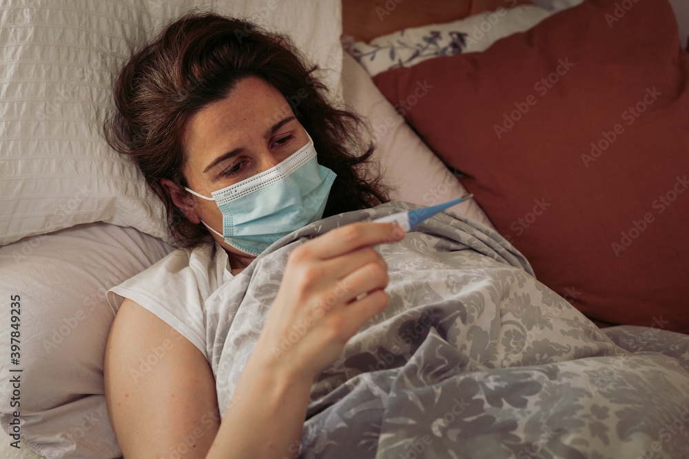 Covid-19 infected woman lying in bed at home in self isolation and checking thermometer. Coronavirus and quarantine concept.  Earthy color tones.
