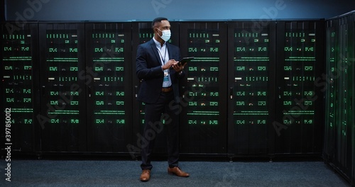 African American handsome man in mask walking among servers with tablet device, tapping on screen and checking big data processors in storage. Male analytic in datacenter working on digital security