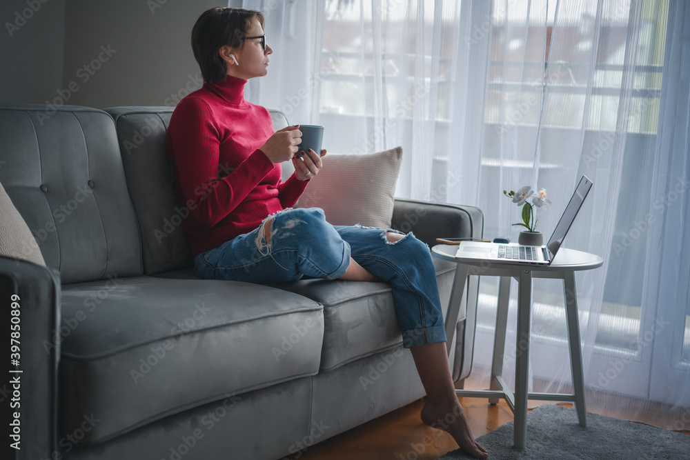 Adult woman working at home online on the phone and laptop with cup of coffee in her hands with a large window in the background Adult woman working at home online on the phone and laptop with cup of coffee in her hands with a large window in the background