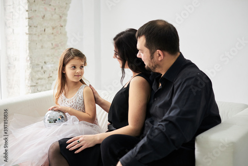Young family of three sitting on white sofa in light room with green plant. Mother, father and daughter, wearing fancy attire relaxing on New Year party. Stay home for winter holidays. Family values.
