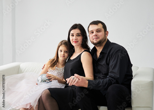 Young family of three sitting on white sofa in light room with green plant. Mother, father and daughter, wearing fancy attire relaxing on New Year party. Stay home for winter holidays. Family values. © Natalia