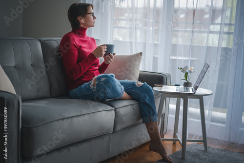 Adult woman working at home online on the phone and laptop with cup of coffee in her hands with a large window in the background