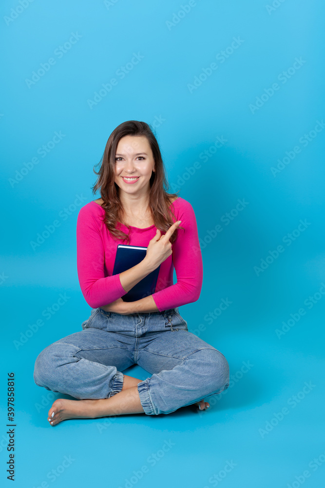 a full-length smiling young woman is sitting on the floor, holding a book and pointing with her index finger at an empty space for text. a full-length smiling young woman is sitting on the floor, holding a book and pointing with her index finger at an empty space for text.