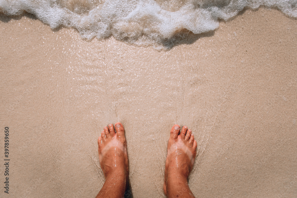 suntanned human feet with stripes of white skin from sandals in summer standing on the sand and sea waves 