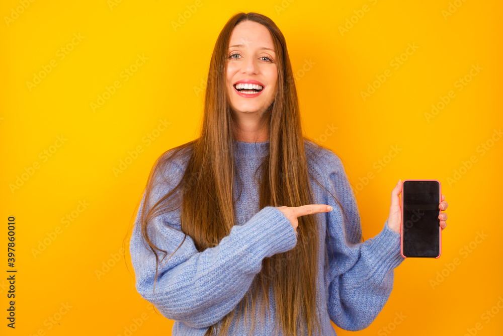 Attractive cheerful Young beautiful Caucasian woman wearing blue sweater against yellow wall holding in hands cell showing black screen Attractive cheerful Young beautiful Caucasian woman wearing blue sweater against yellow wall holding in hands cell showing black screen
