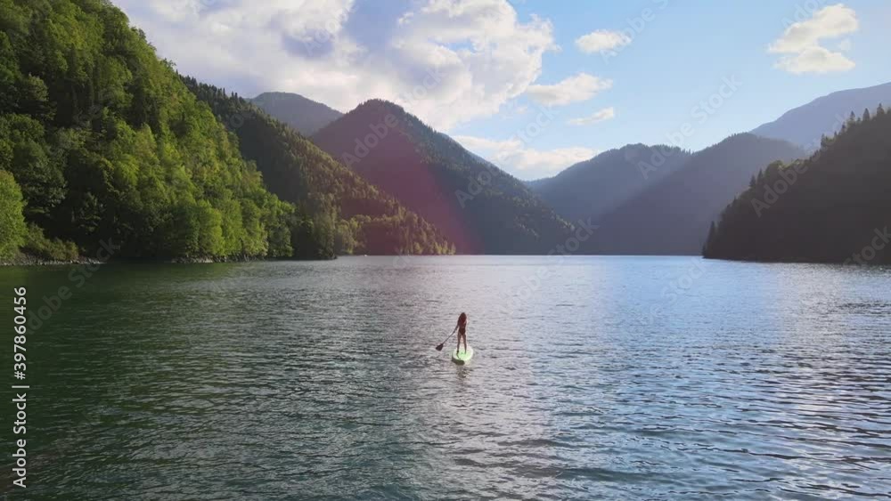 Aerial drone view of pretty sporty woman paddle on sup board at mountain lake during sunset. Female silhouette doing water sport during summer holidays. Concept of active tourism and travel. Aerial drone view of pretty sporty woman paddle on sup board at mountain lake during sunset. Female silhouette doing water sport during summer holidays. Concept of active tourism and travel.