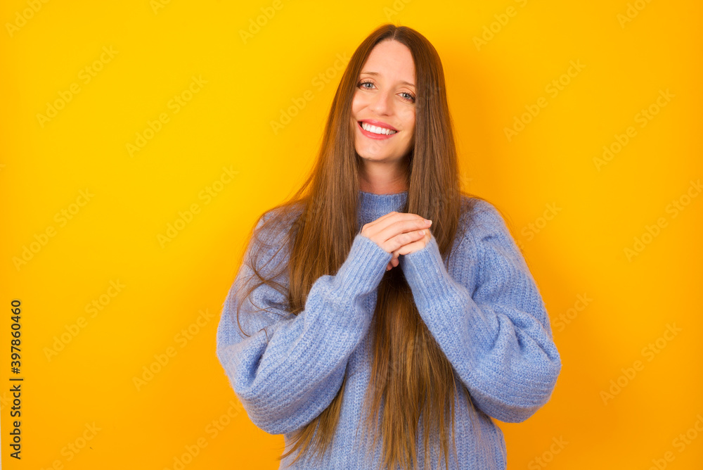 Positive Young beautiful Caucasian woman wearing blue sweater against yellow wall smiles happily, glad to receive pleasant news from interlocutor, keeps hands together. People emotions concept. Positive Young beautiful Caucasian woman wearing blue sweater against yellow wall smiles happily, glad to receive pleasant news from interlocutor, keeps hands together. People emotions concept.