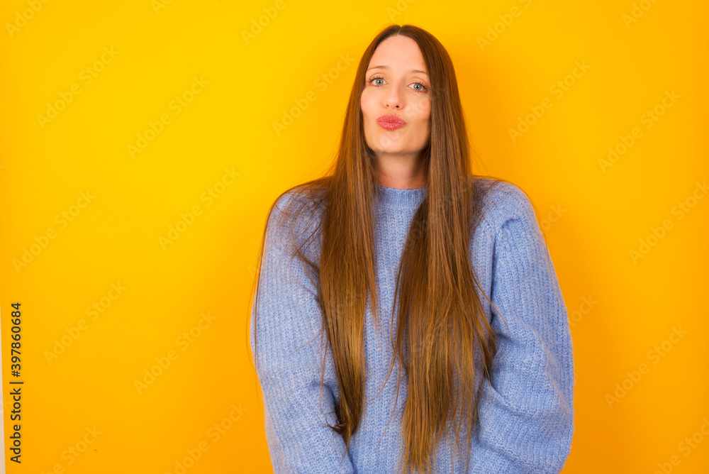 Shot of pleasant looking Young beautiful Caucasian woman wearing blue sweater against yellow wall, pouts lips, looks at camera, Human facial expressions Shot of pleasant looking Young beautiful Caucasian woman wearing blue sweater against yellow wall, pouts lips, looks at camera, Human facial expressions