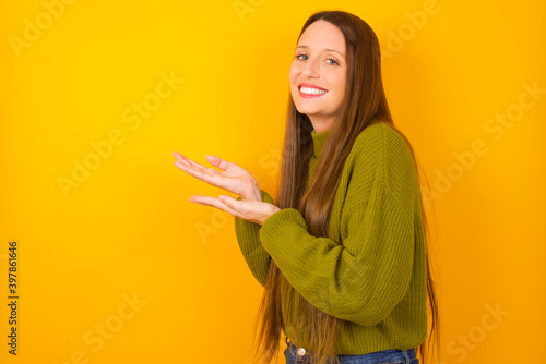 Young beautiful Caucasian woman wearing green sweater against yellow wall pointing aside with hands open palms showing copy space, presenting advertisement smiling excited happy
