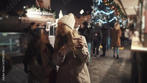 Pretty young girl surprising friend, running into her from behind. Teen friends standing in a middle of christmas fare, drinking coffee, having great time together.