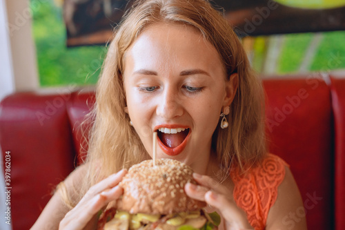 Preety and cute woman with blond hair is bitting a huge burger in a cafe. Fast food concept. Portrait.