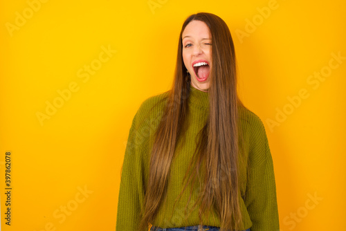 Young beautiful Caucasian woman wearing green sweater against yellow wall winking looking at the camera with sexy expression, cheerful and happy face. © Roquillo