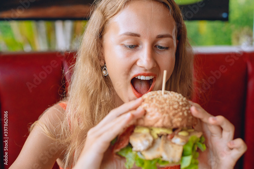Young beautiful lady with blond hair is bitting a huge burger in a cafe. Fast food concept. Portrait.