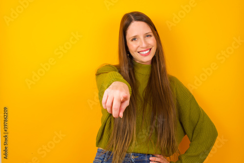 Young beautiful Caucasian woman wearing green sweater against yellow wall pointing at camera with a satisfied, confident, friendly smile, choosing you