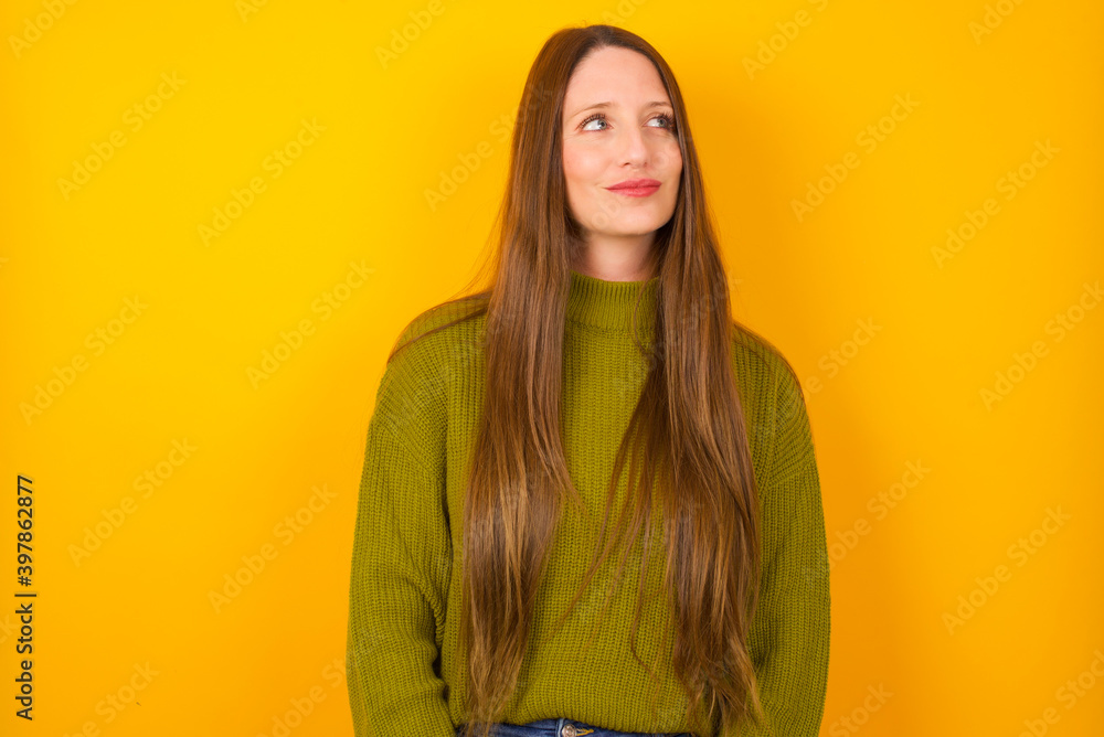 Young beautiful Caucasian woman wearing green sweater against yellow wall looking aside into empty space thoughtful