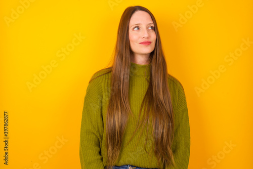 Young beautiful Caucasian woman wearing green sweater against yellow wall looking aside into empty space thoughtful © Roquillo
