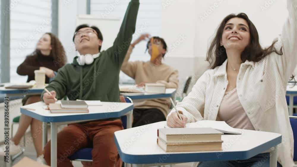 Diverse students sitting by desks in classroom, writing in notebooks, taking off colorful face masks, throwing them up, Mixed-Race girl looking, smiling on camera Diverse students sitting by desks in classroom, writing in notebooks, taking off colorful face masks, throwing them up, Mixed-Race girl looking, smiling on camera