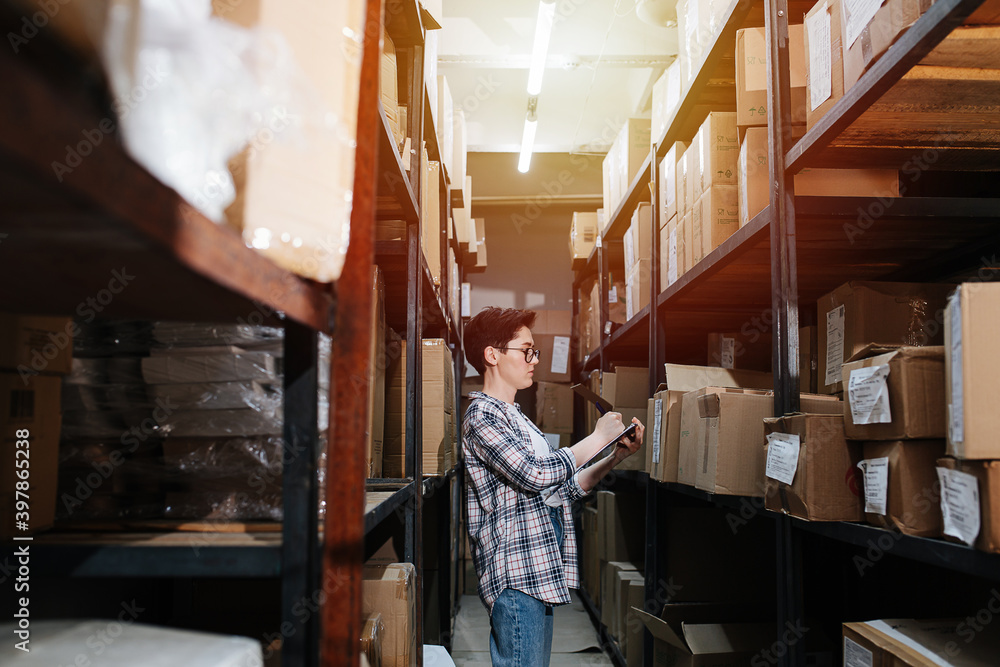 Short-haired woman taking list of warehouse inventory. Side view. Tall shelves with boxes and tags on both side of her. She wears sheckered shirt and glasses.