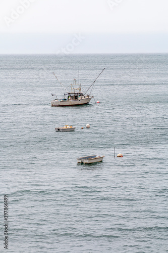 A fishing trawler is at anchor in a ocean bay. It has to poles out on each side of the boat. A buoy is between the boat and a small dingy. The water is grey. © Timothy
