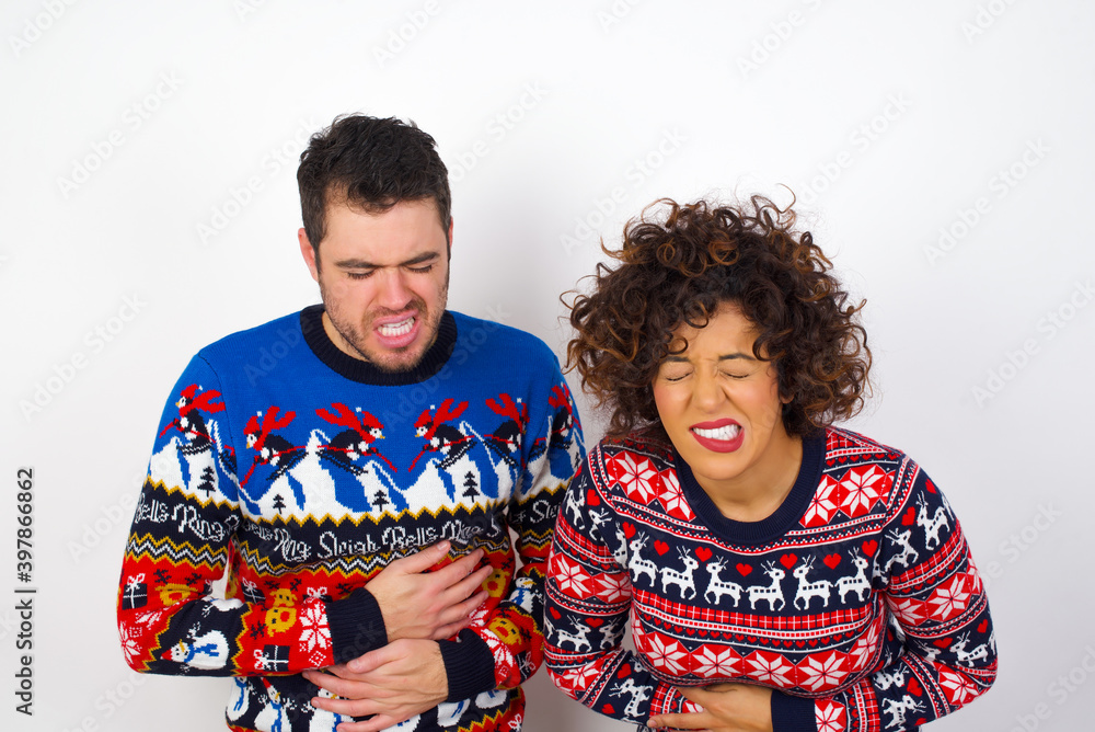 Young couple wearing Christmas sweater standing against white wall with hand on stomach because nausea, painful disease feeling unwell. Ache concept.
