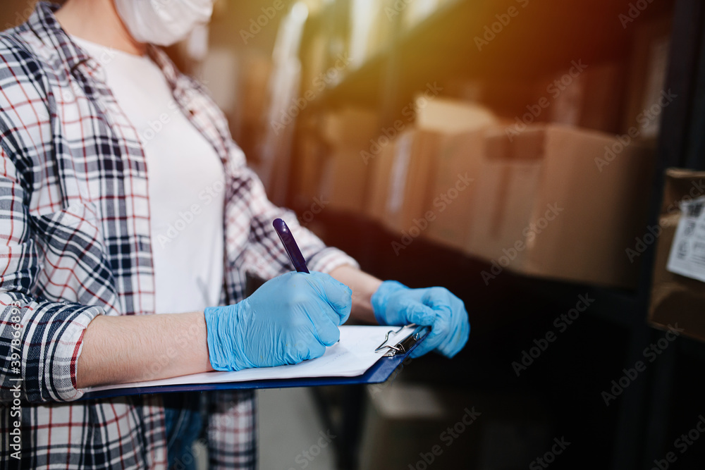 Cropped image of a warehouse worker filling in list on a pad. She's standing next to a tall shelf with boxes. Wearing blue gloves.